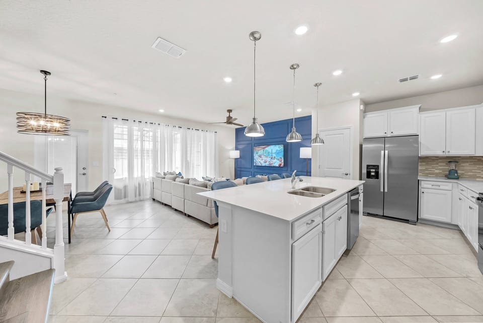This bright kitchen and living area features sleek white cabinetry and a vibrant blue accent wall. The open layout, complemented by modern lighting and cozy seating, invites you to relax and socialize comfortably.