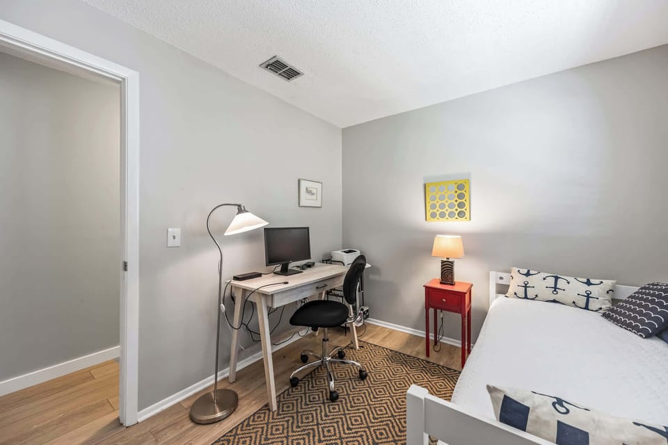 Compact home office area with a desk, computer, and task lighting next to a bed with nautical-themed pillows.