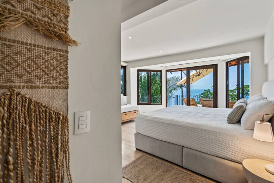 Sunlit second bedroom opens to tropical treetops and azure ocean horizons at Casa Tiempo Puerto Vallarta. Natural textures and crisp linens create a peaceful retreat in this luxury palapa villa near Playa Conchas Chinas.