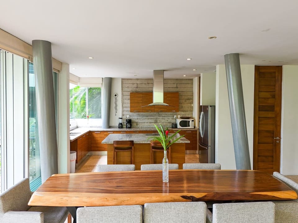 Live-edge wood dining table with comfy chairs beside an open kitchen with breakfast bar, granite counters and stainless appliances. Bright, airy space for family dinners or remote work in Puerto Vallarta, 5 min to Zona Romántica & Conchas Chinas.