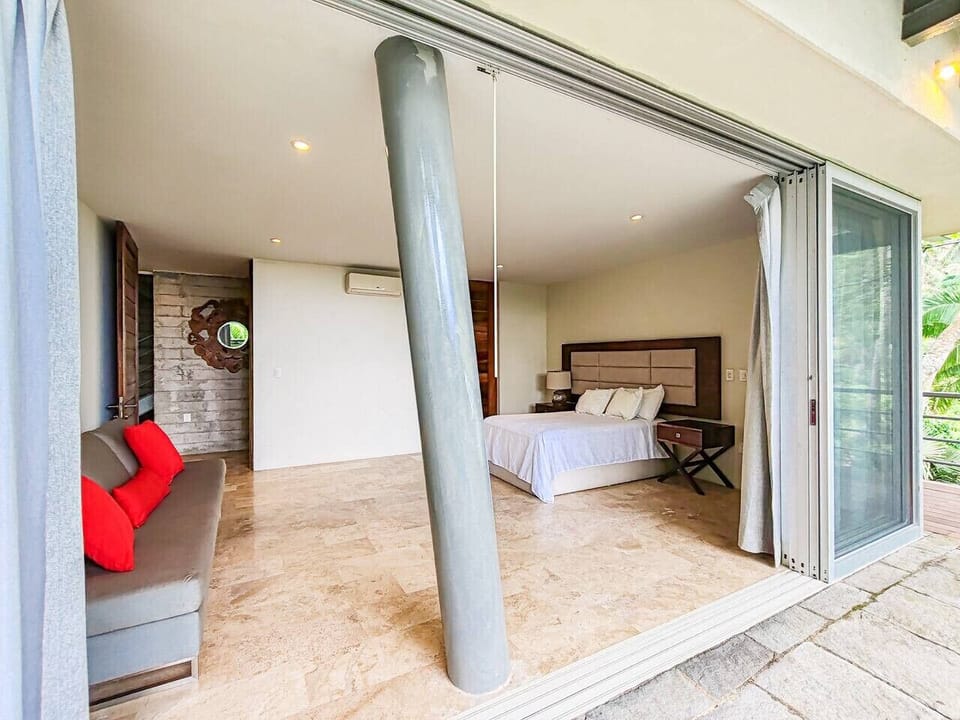 Bedroom 3 is a spacious suite with king bed, A/C, marble floors and a cozy sofa for reading or an extra lounge spot. Large sliding doors open to the pool deck and jungle views, creating a breezy indoor-outdoor feel and easy access for morning dips.