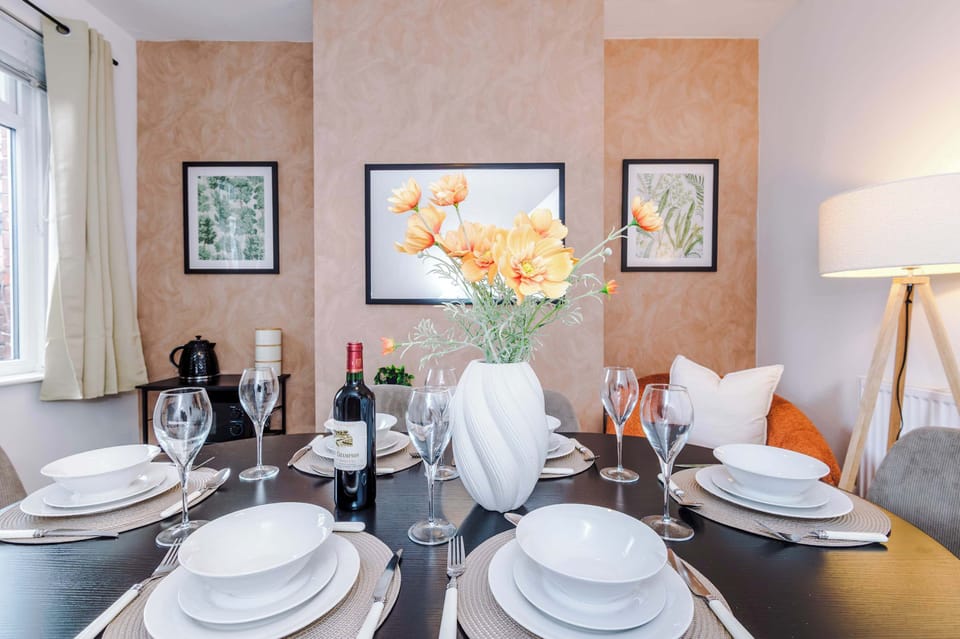 A modern dining space set for six, with a sleek oval table, fresh flowers at the center, and elegant place settings. Paired with a bold orange sofa and framed art, the room blends warmth and sophistication.