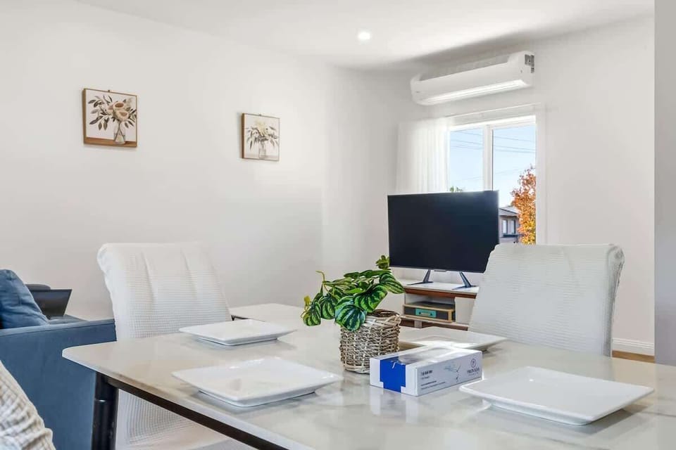 Bright Modern Dining This crisp, sunlit Airbnb space features a set dining table with high-back white chairs, leading seamlessly into the lounge zone. Air conditioning and minimalist decor ensure a breezy, comfortable stay.