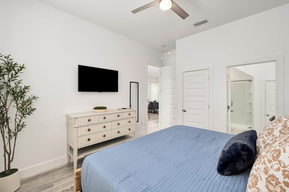 Unwind in your serene bedroom featuring soft linens, a calming blue quilt, and a mounted TV for relaxation. The ceiling fan keeps you cool while the bright, airy space invites restful sleep.