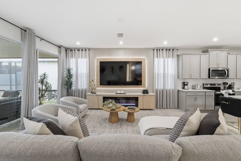 Reúna-se em uma sala integrada e iluminada, com sofá confortável, TV grande, lareira elétrica e cozinha ao lado. Tons neutros, cortinas altas e acesso à piscina criam um ambiente acolhedor.