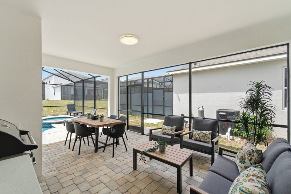 Relax in a covered screened patio with cozy outdoor seating, a wood-top coffee table, colorful pillows, and sliding doors connecting to the home. A bright, comfortable spot to enjoy fresh air and quiet moments.