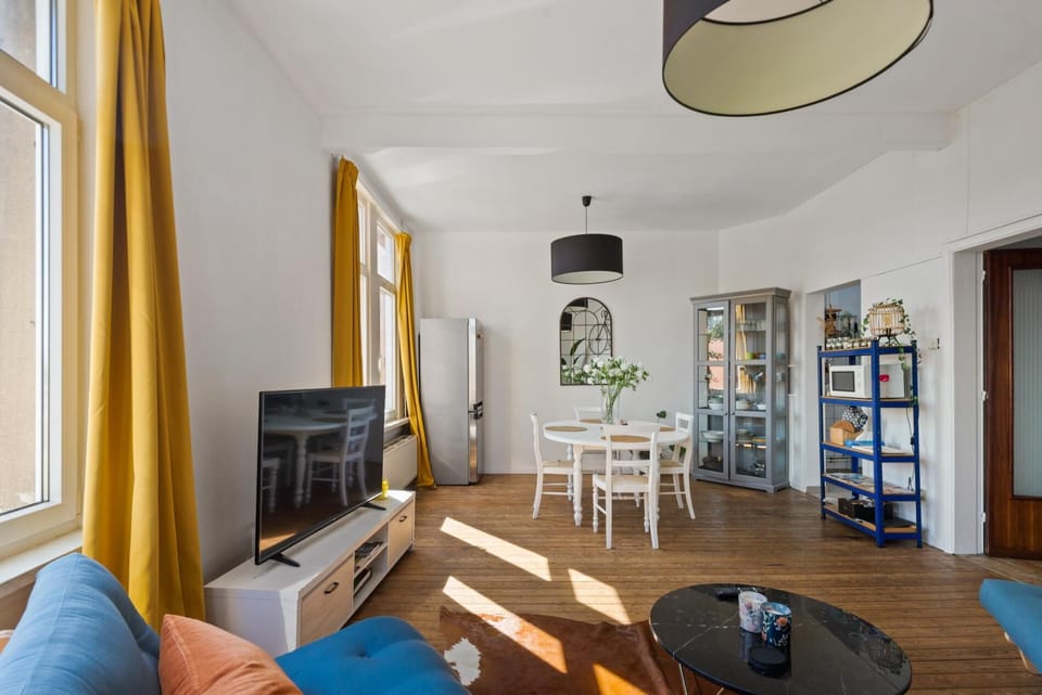 Open living and dining area with tall windows and warm natural light throughout the space. The comfortable seating area, TV, and dining table create a relaxed setting to unwind, share meals, or enjoy quiet moments indoors.