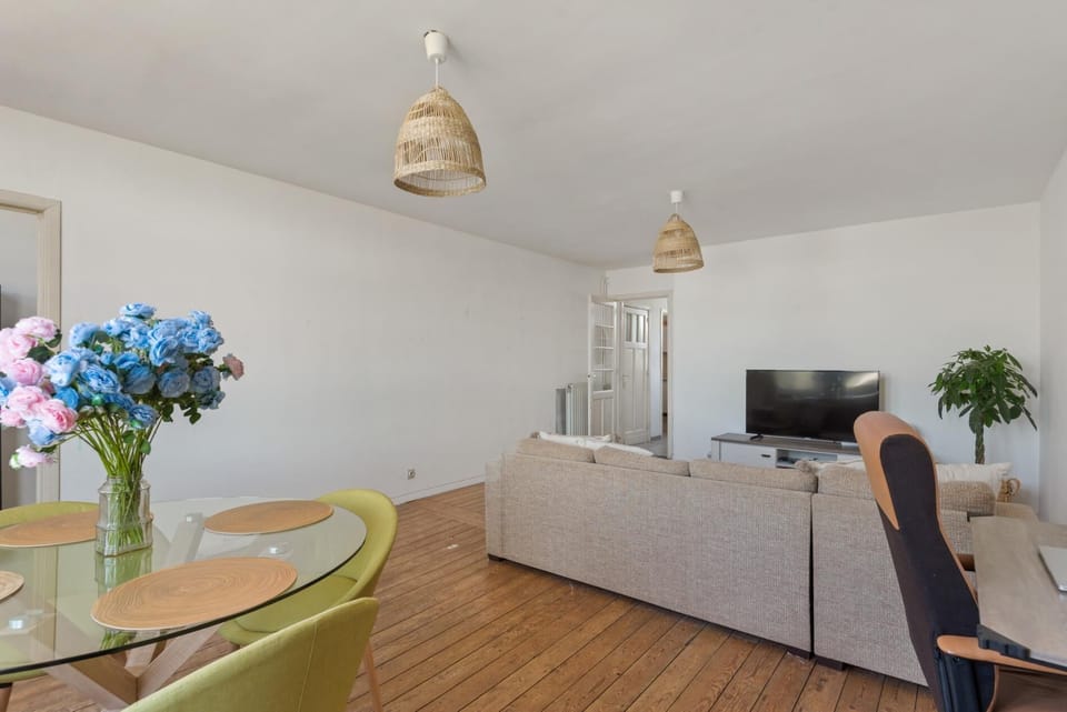 Spacious open living and dining area with wooden floors, comfortable seating, and a TV for relaxing evenings. Large windows bring in natural light, while the dining table offers a pleasant space to gather or enjoy a quiet meal.