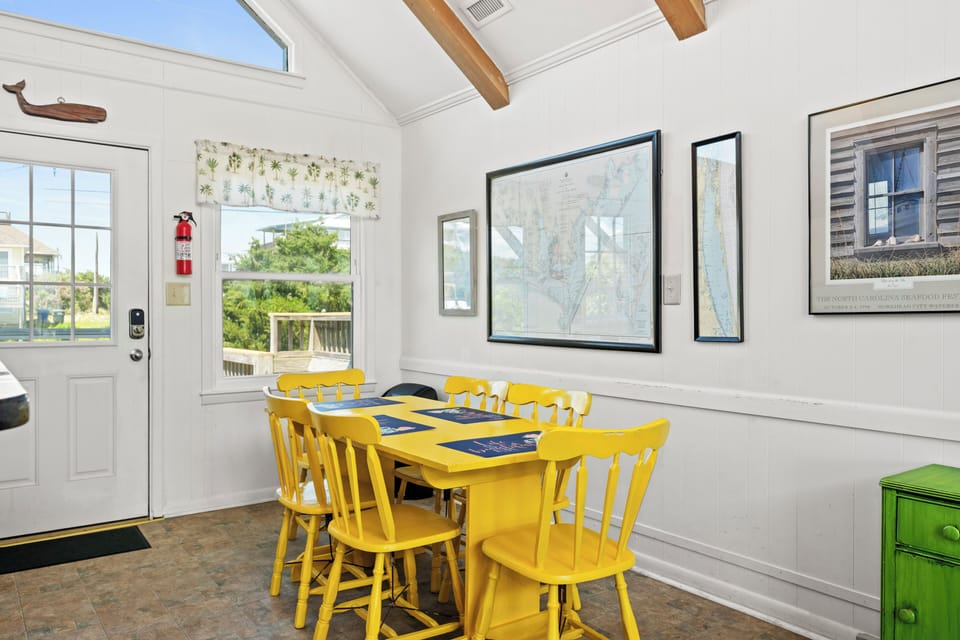 Gather around this cheerful yellow dining table with vaulted ceilings, warm wood beams, and natural light streaming through coastal-inspired windows—your perfect spot for shared meals and vacation memories.