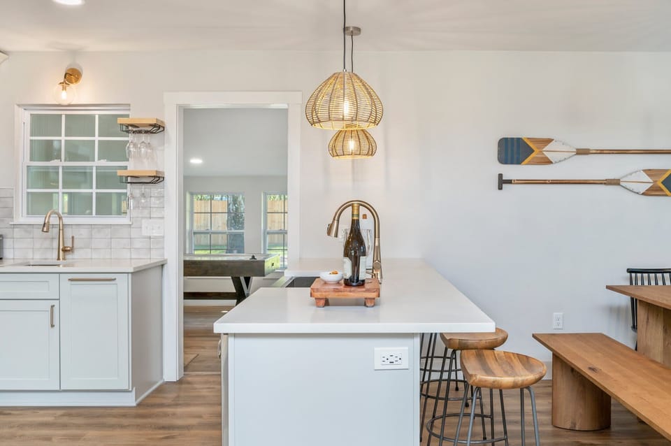 Enjoy this bright and airy perspective of the kitchen and dining area. With its sleek white countertops, coastal-themed wall decor, and warm wicker lighting, this open space offers a seamless flow for entertaining and shared meals.