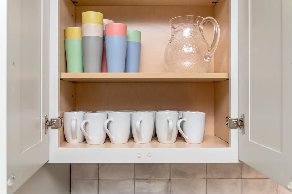 Find everything you need for your morning brew or a refreshing drink! This neatly organized cabinet is stocked with a set of clean white mugs, colorful tumblers, and a glass pitcher, ensuring you're fully equipped for your stay.