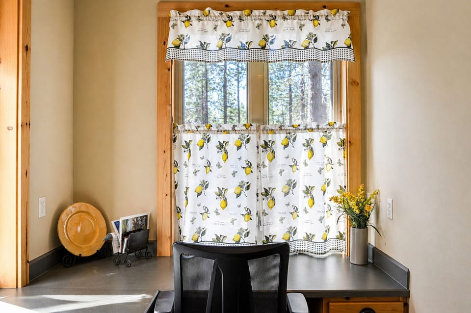 Your charming kitchen nook features cheerful lemon-print curtains framing forest views, creating the perfect spot for morning coffee.
