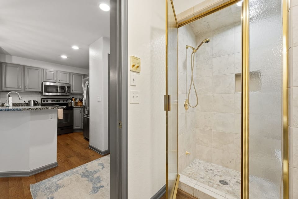 Walk-in shower with brass trim, situated next to a view of the kitchen.
