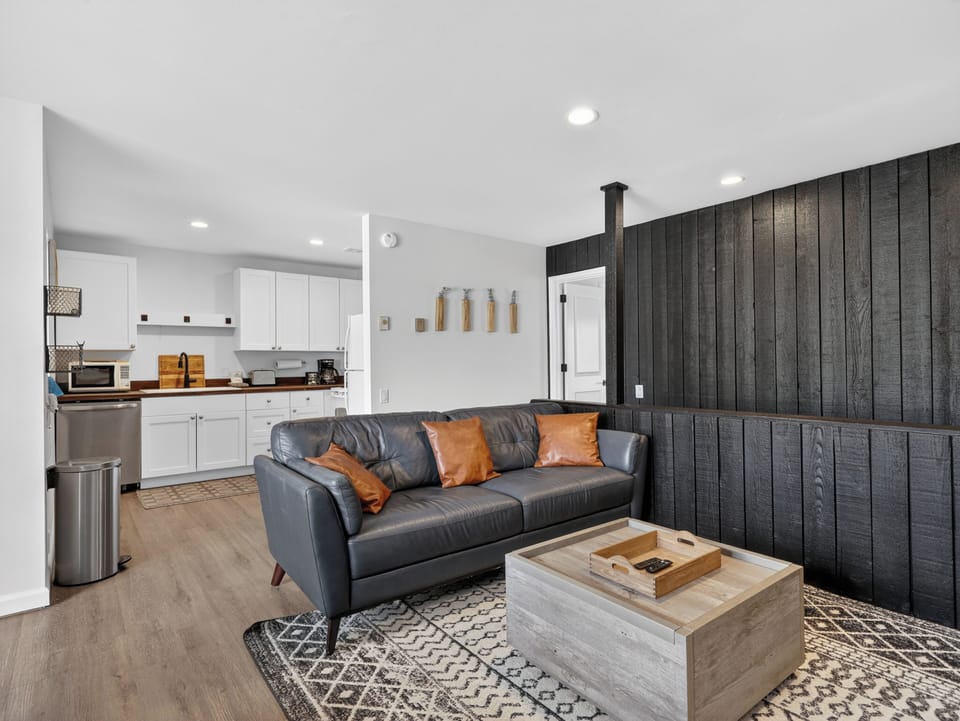 Sink into the leather sofa and take in the striking dark wood accent wall, complemented by warm orange throw pillows. The open layout connects you to a bright, fully-equipped kitchen, perfect for your stay.
