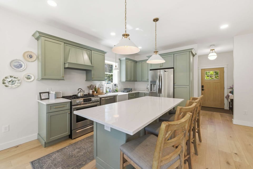 The kitchen island seats four and anchors the whole main level — sage green cabinetry, brass hardware, and wide-plank hardwood floors throughout.