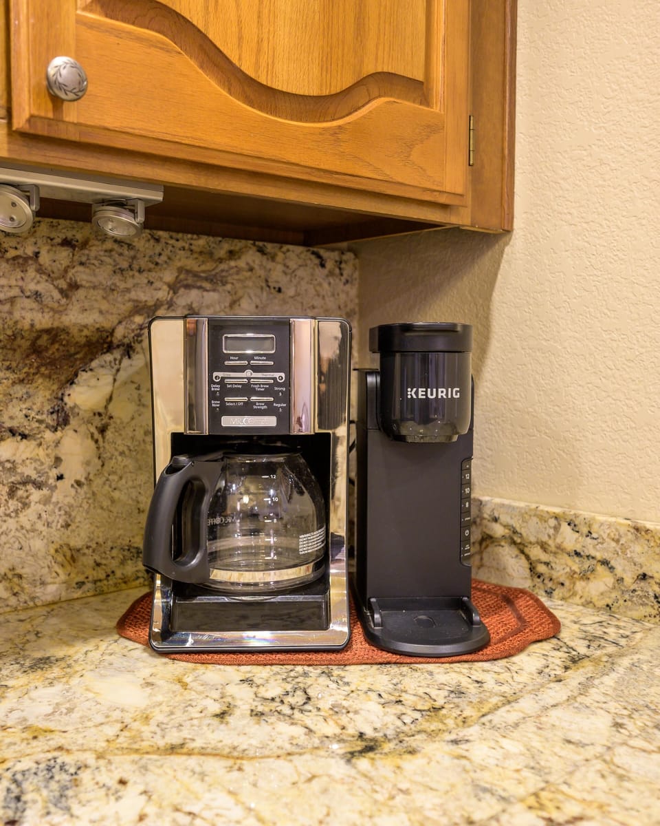 A dual coffee maker setup sits on a speckled granite countertop beneath warm wood cabinetry.