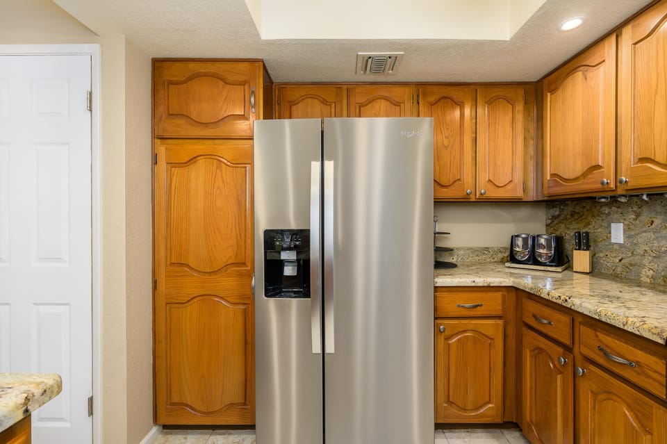 Stainless steel side-by-side refrigerator stands between honey oak cabinets and granite countertops in the kitchen.