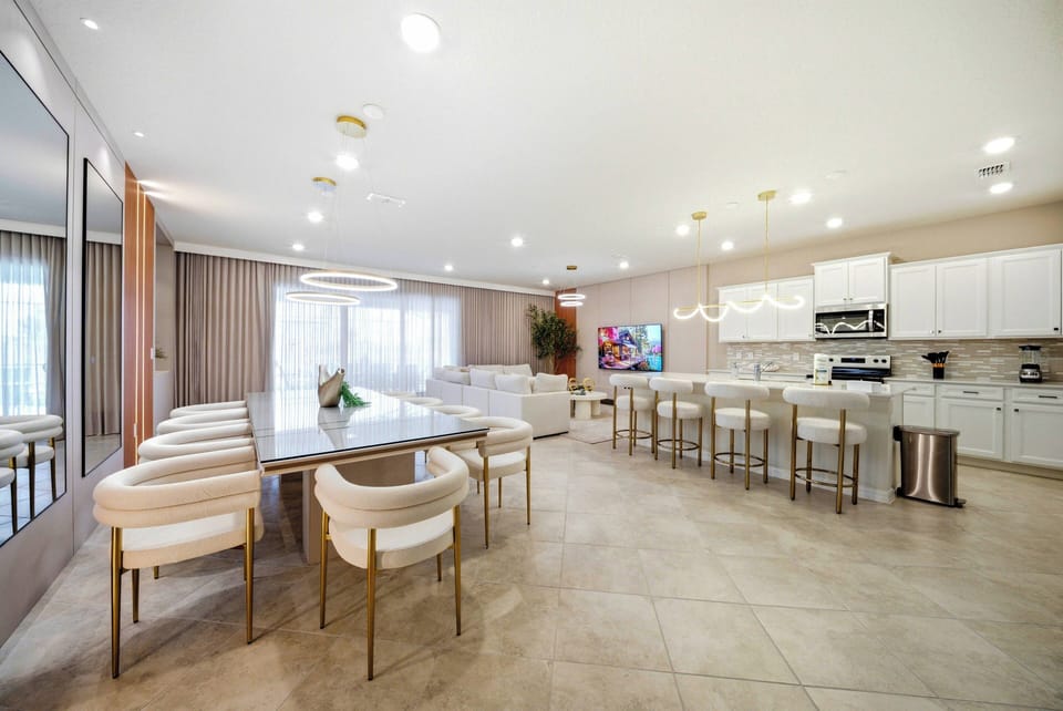 This bright, spacious kitchen features sleek white cabinetry and a large dining table with stylish upholstered chairs, ideal for gatherings. Expansive windows let in natural light, enhancing the inviting, modern atmosphere.