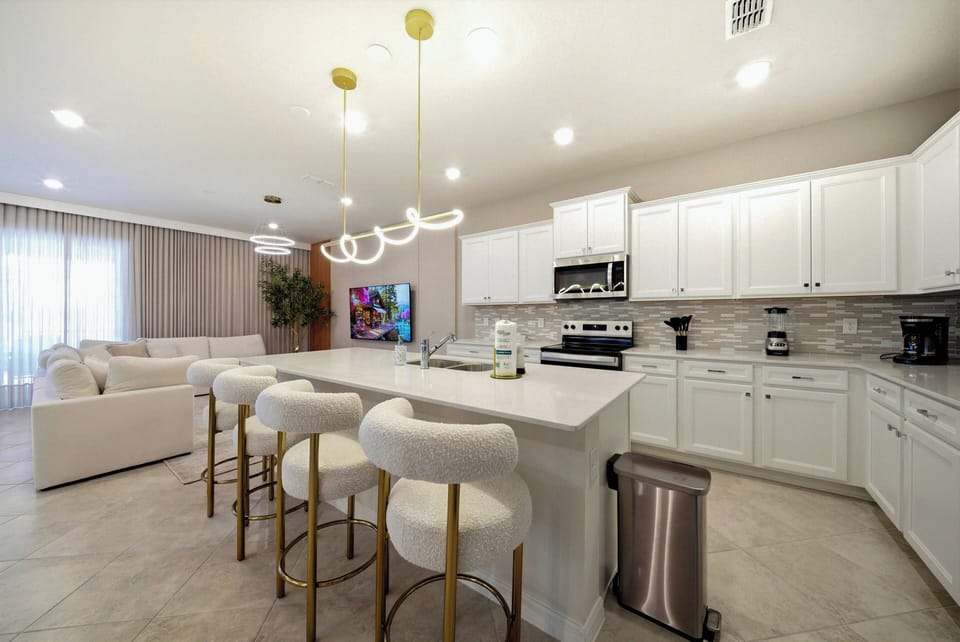 This bright kitchen features sleek white cabinetry with elegant golden accents, creating a sophisticated vibe. The spacious island with comfortable seating is perfect for casual dining, while large windows bathe the room in.