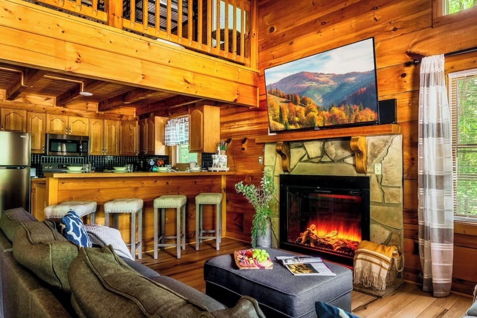 Great room overview showing the open-concept layout with kitchen bar, roaring fireplace, flat-screen TV, and loft visible above