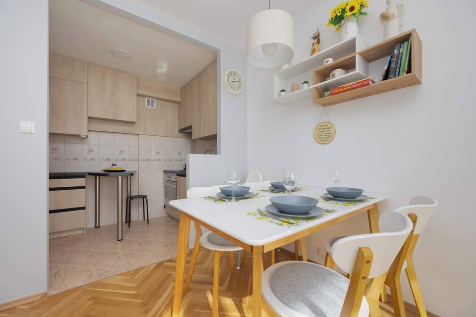A dining area features a white table paired with four wooden chairs, set within a bright, open-plan space. Decorative wall shelving and a pendant light add a homely touch near the kitchen entrance.