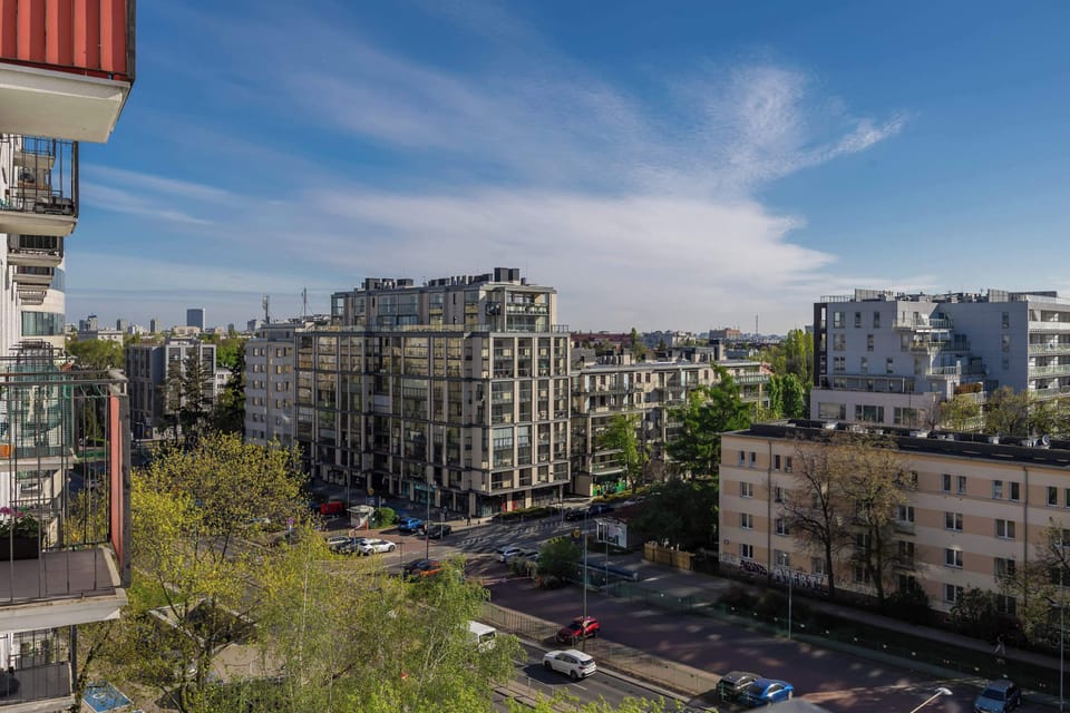 The balcony overlooks a busy city street lined with diverse architecture and mature trees. The wide perspective highlights the vibrant urban environment under a clear sky.