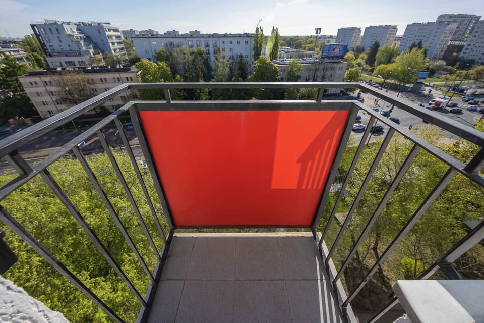 A private balcony with a red privacy screen offers a view of the surrounding trees and residential buildings. The elevated position provides a quiet outdoor spot to enjoy the urban landscape.
