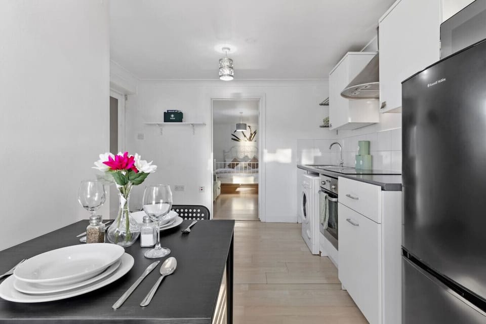 Set dining table for two beside a compact white kitchenette, with a black fridge and a view through to the bedroom beyond.