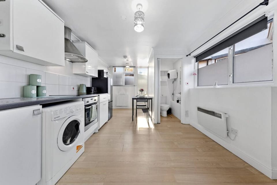 Bright galley kitchen with white cabinets, washing machine and oven, black fridge, and a small dining table beside a window, with a compact WC visible through the open doorway.