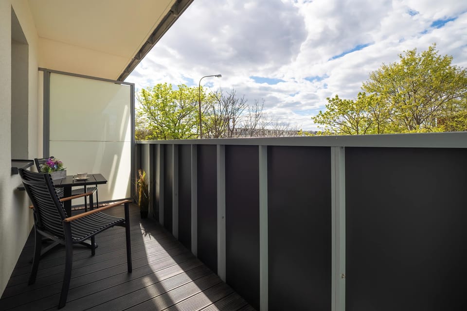 A narrow balcony with dark railings and a small seating setup overlooking a bright outdoor view.