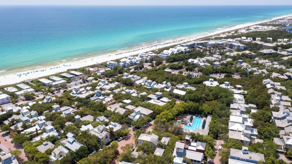 Aerial view of a coastal vacation community featuring white beach houses nestled among lush greenery, steps from pristine turquoise waters and sandy shores.