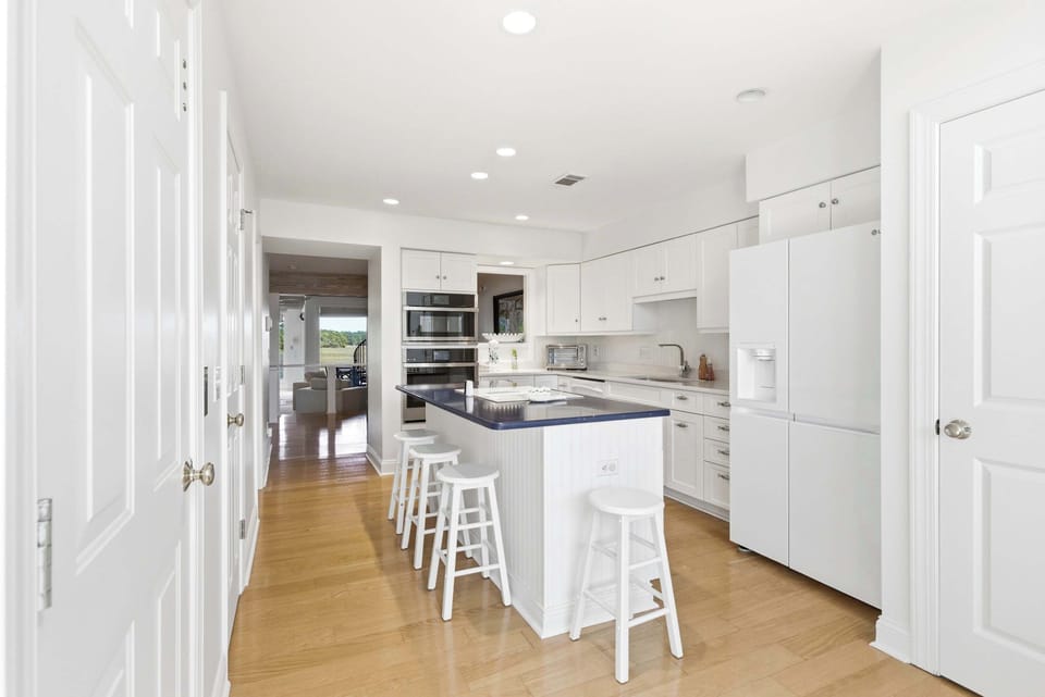 Full kitchen with double wall ovens, a large navy island with bar stools, and a hallway sightline straight to the water views beyond.