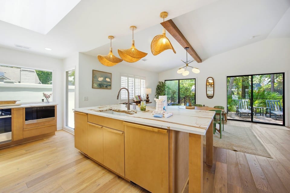 Woven wicker pendants cast a soft glow over the marble island and warm wood cabinetry. High ceilings and wide glass doors pull the lush greenery indoors, creating a bright and airy culinary space perfect for slow mornings.