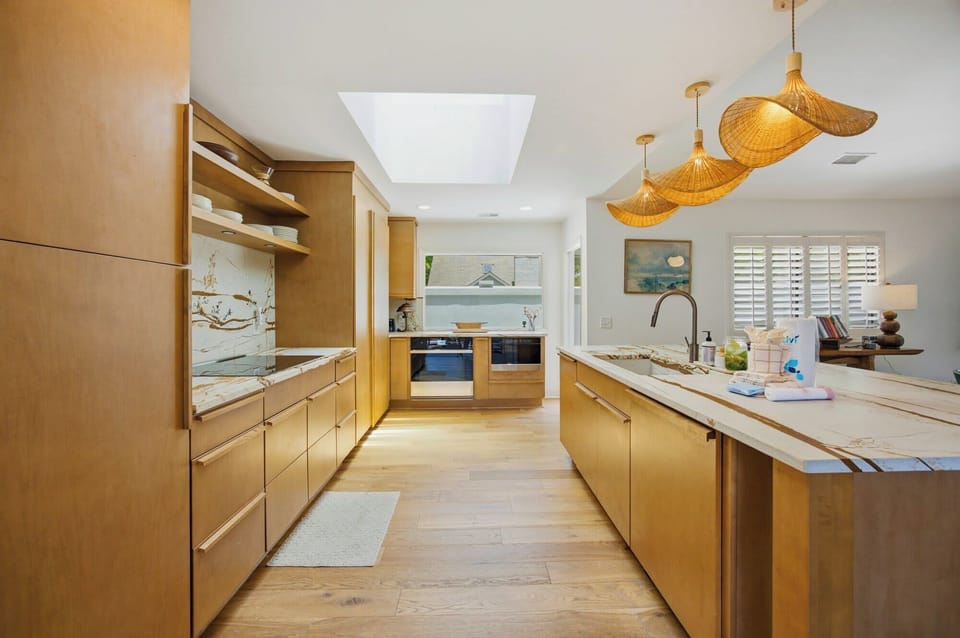 Amber wood cabinetry and marble counters glow beneath a bright skylight, creating a serene culinary space. Rattan pendants add a rhythmic texture over the island, perfect for slow mornings and shared meals in this airy kitchen.