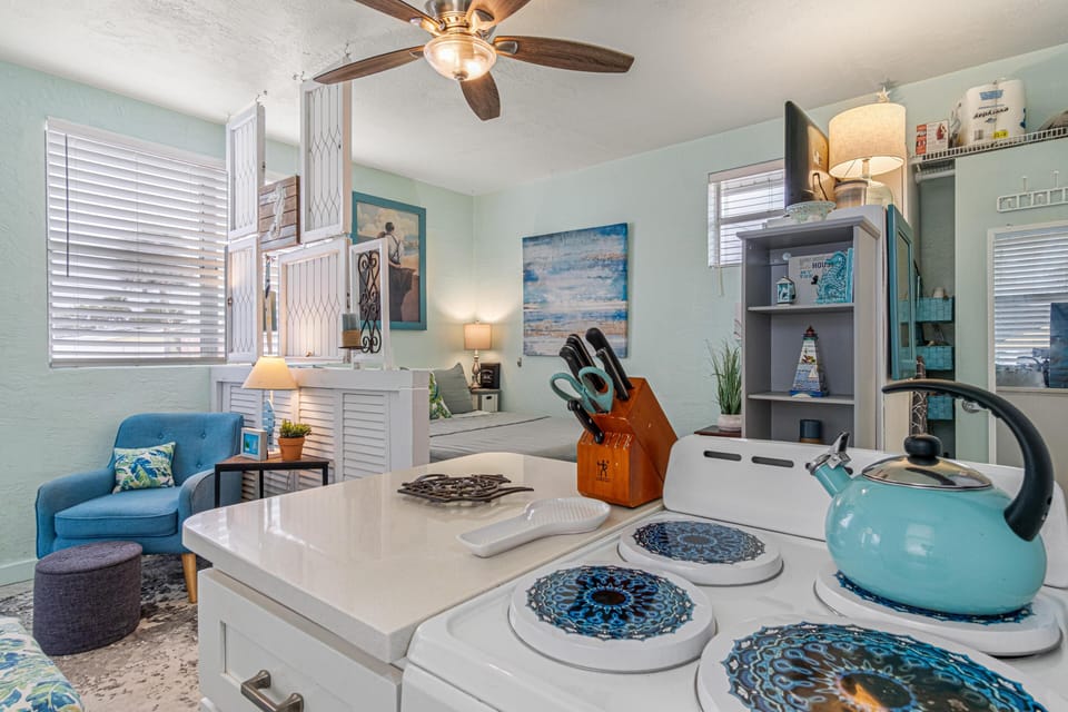 The kitchen stove with blue patterned burner covers faces a soft blue armchair and a bed separated by white wooden shutters.