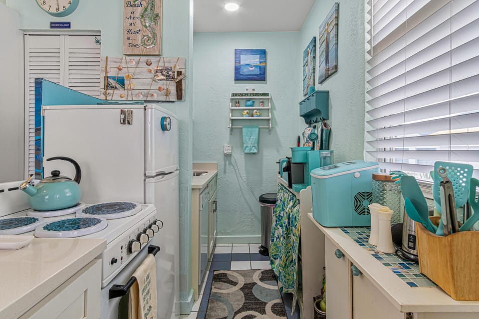 Narrow kitchen with white appliances, turquoise kettle, blue-patterned burner covers, and coastal-themed wall decor.