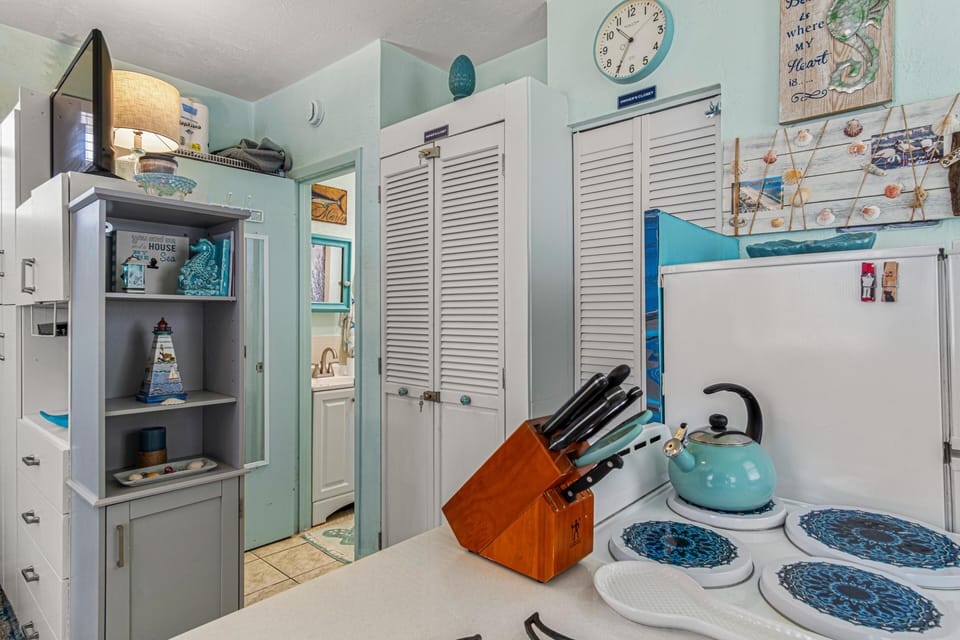 A compact kitchen corner with white appliances, turquoise kettle, and louvered pantry doors under a round wall clock.