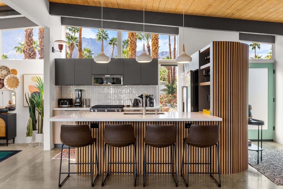 Your sleek modern kitchen features premium appliances and a stunning breakfast bar, all framed by desert palms through floor-to-ceiling windows.