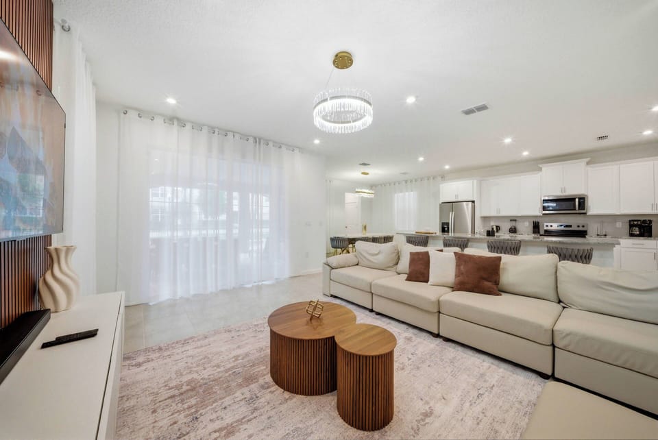This bright and airy living room features a spacious sectional sofa that invites relaxation, warm wooden coffee tables, and large windows with sheer curtains that fill the space with natural light for a welcoming atmosphere.