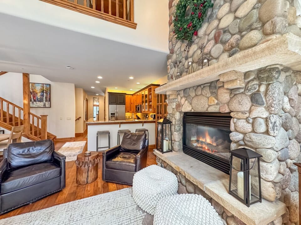 River rock rises around a glowing gas fireplace, anchoring a space where kitchen warmth, dining laughter, and living room ease all flow together naturally