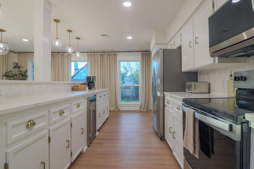 Remodeled kitchen with new electric range, stainless fridge, wine cooler, dual coffee maker, toaster, and ample cabinet storage. Gold hardware finishes and natural light from a large window. Everything a family needs to cook comfortably.