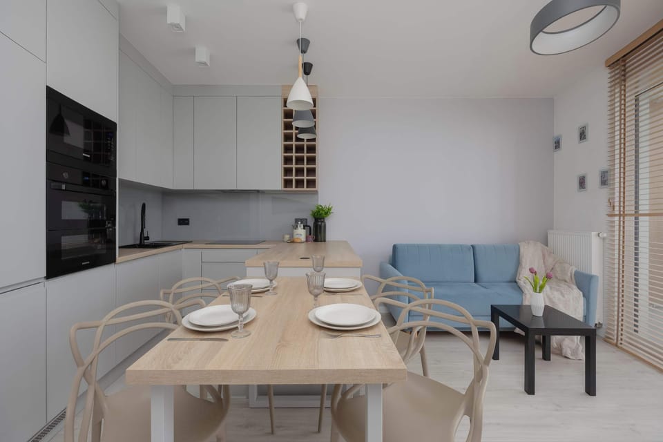 An open-plan living space with a dining table, kitchenette, and blue sofa. Bright windows and modern finishes give the apartment a welcoming feel.