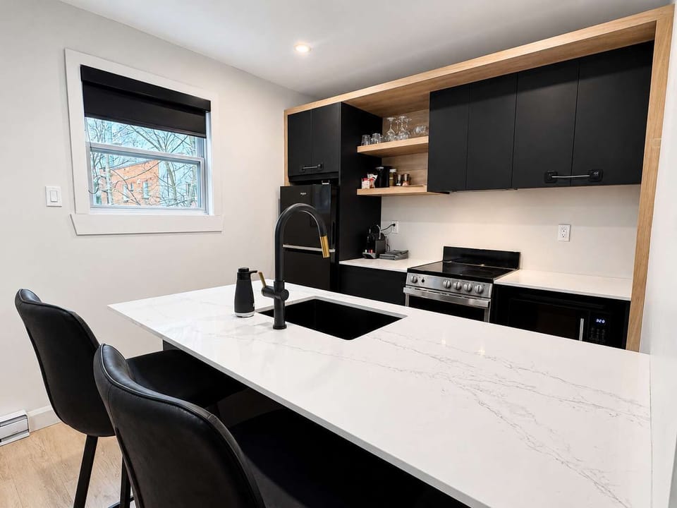 Une cuisine minimaliste contemporaine comprenant un comptoir d'îlot blanc marbré équipé d'un évier noir intégré, d'un robinet noir élégant et de tabourets de bar en cuir noir épurés.