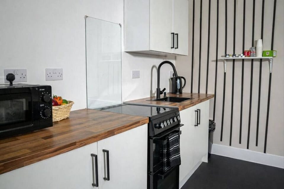 Kitchenette with microwave, hob and oven, sink, kettle, and wall shelving.