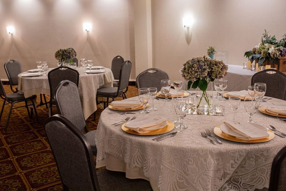 Banquet hall with lace-covered round tables, full place settings, and flower centrepieces under warm wall lighting.