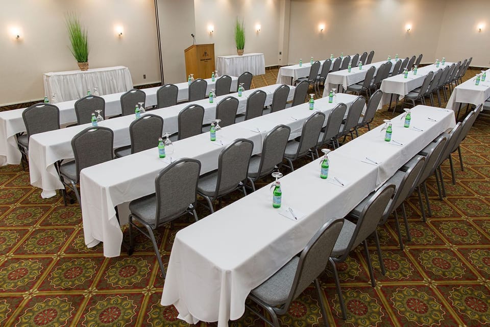 Banquet hall set classroom-style with long white-clothed tables, grey chairs, water bottles, and a podium at the front.