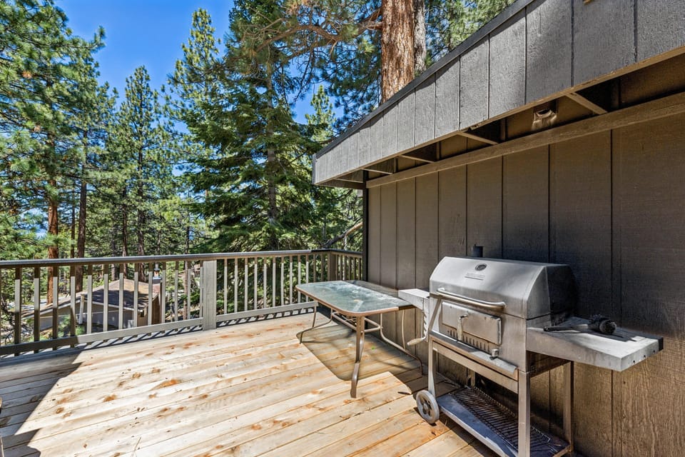 Wood deck with metal grill and folding glass-top table, surrounded by tall pine trees under clear skies.