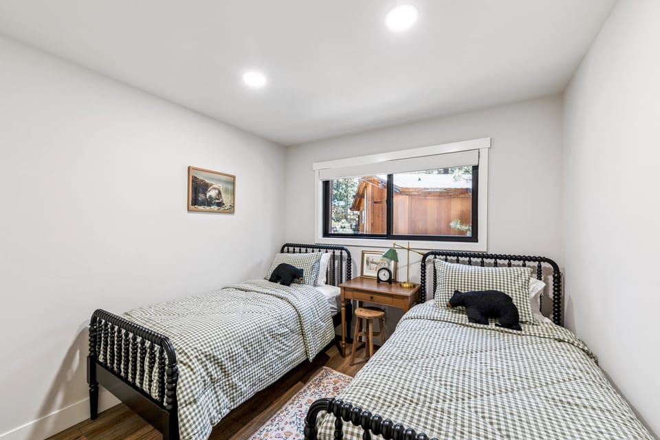 Twin beds with black spindle frames are dressed in green gingham bedding, separated by a wooden nightstand under a window.