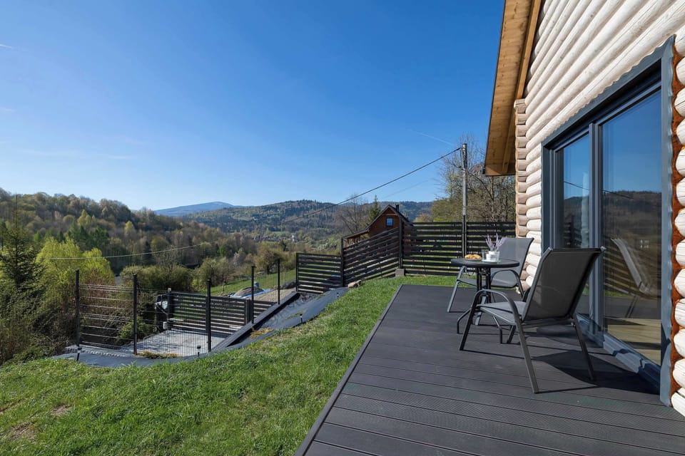 A view of the cottage from the entrance, showing the landing and glass doors. The surroundings are green, and the building blends perfectly with the landscape.