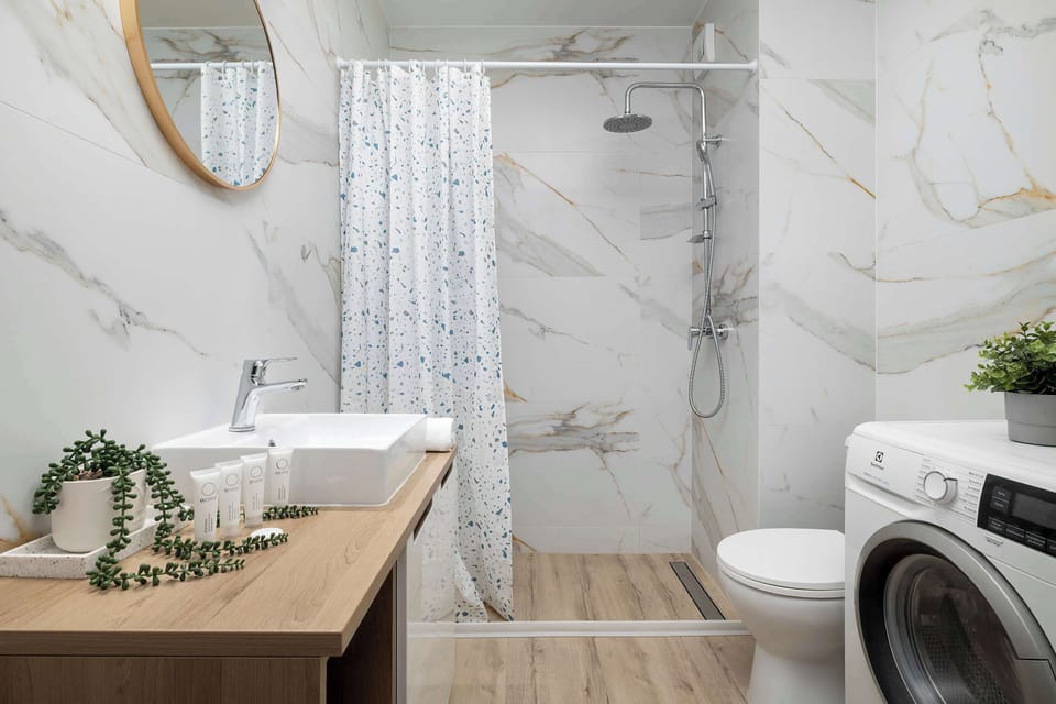 The modern bathroom is styled with marble-patterned tiles and wooden accents. It includes a walk-in shower, a washbasin, and a washing machine.